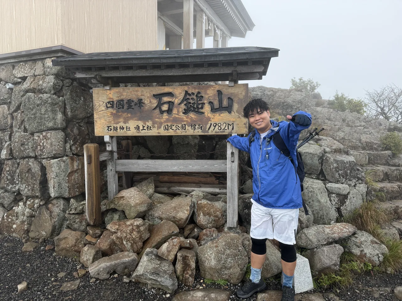 石鎚山頂上社の山名板(標高1982m)の横で親指を立てる迫佑樹