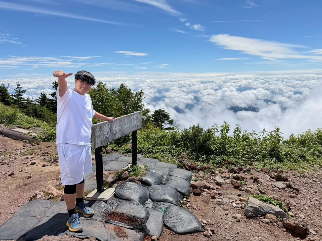 雲海を背に、男体山頂の山名板の横で親指を立てる迫佑樹