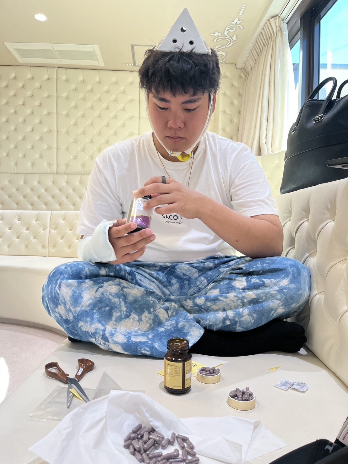 白いキルティングのソファ席で、ピラミッドを頭にかぶり「SACOIN」Tシャツ姿でサプリメントのカプセルを瓶に詰める迫佑樹。