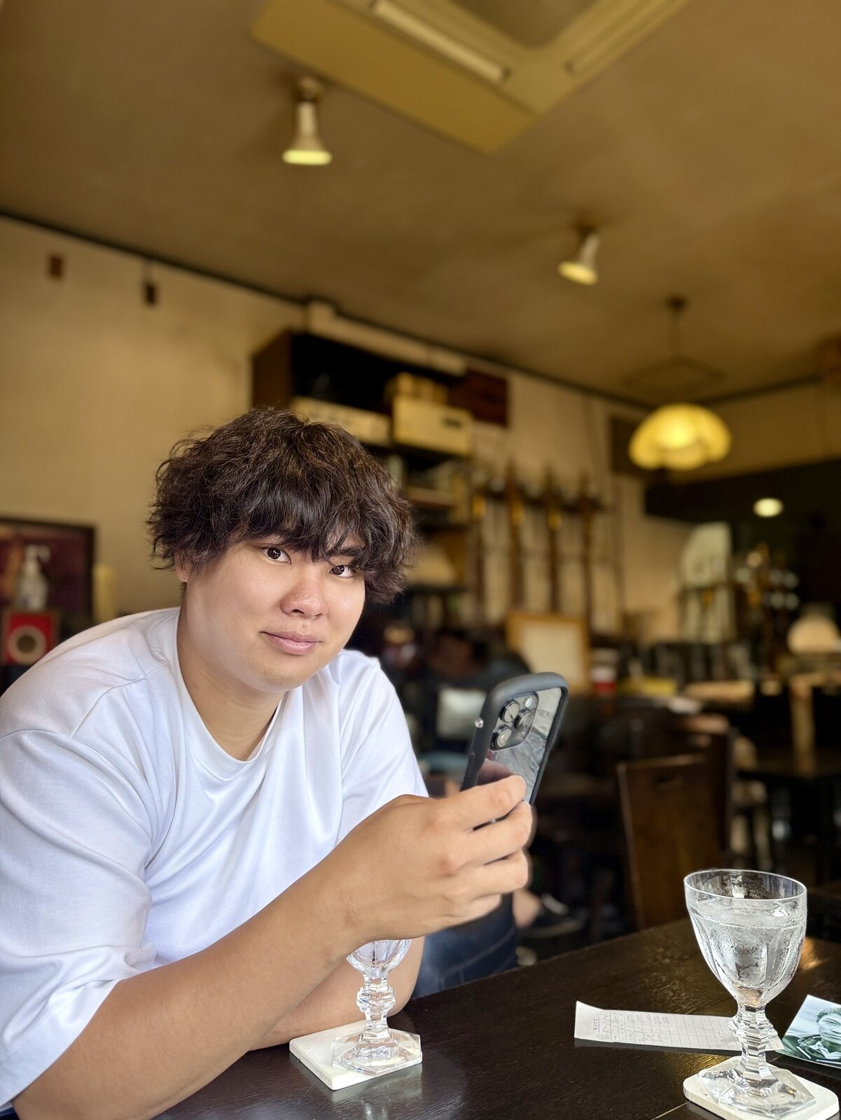 レトロな純喫茶の席で、白いTシャツを着てスマートフォンを手にカメラへ視線を向ける迫佑樹。テーブルにガラスのゴブレット。