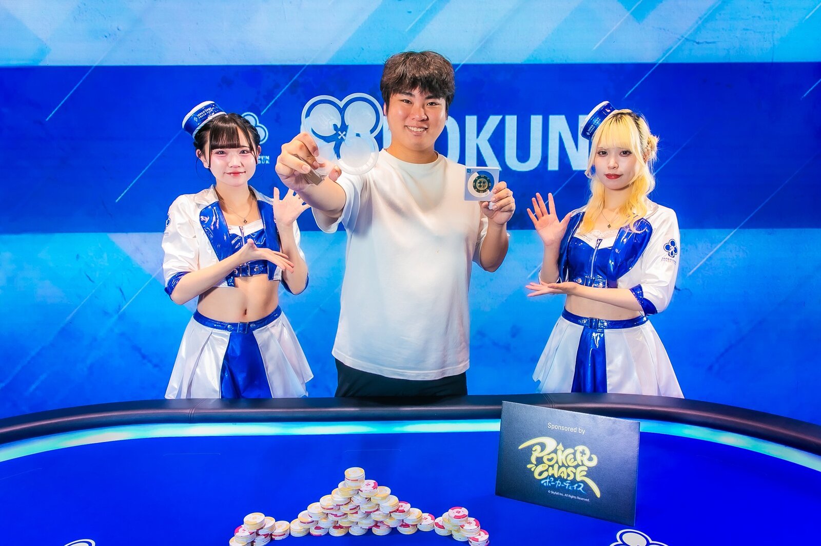 青いステージ上で、優勝メダルをカメラに掲げて笑顔を見せる迫佑樹。両脇にイベントコンパニオン、手前に積まれたポーカーチップ。
